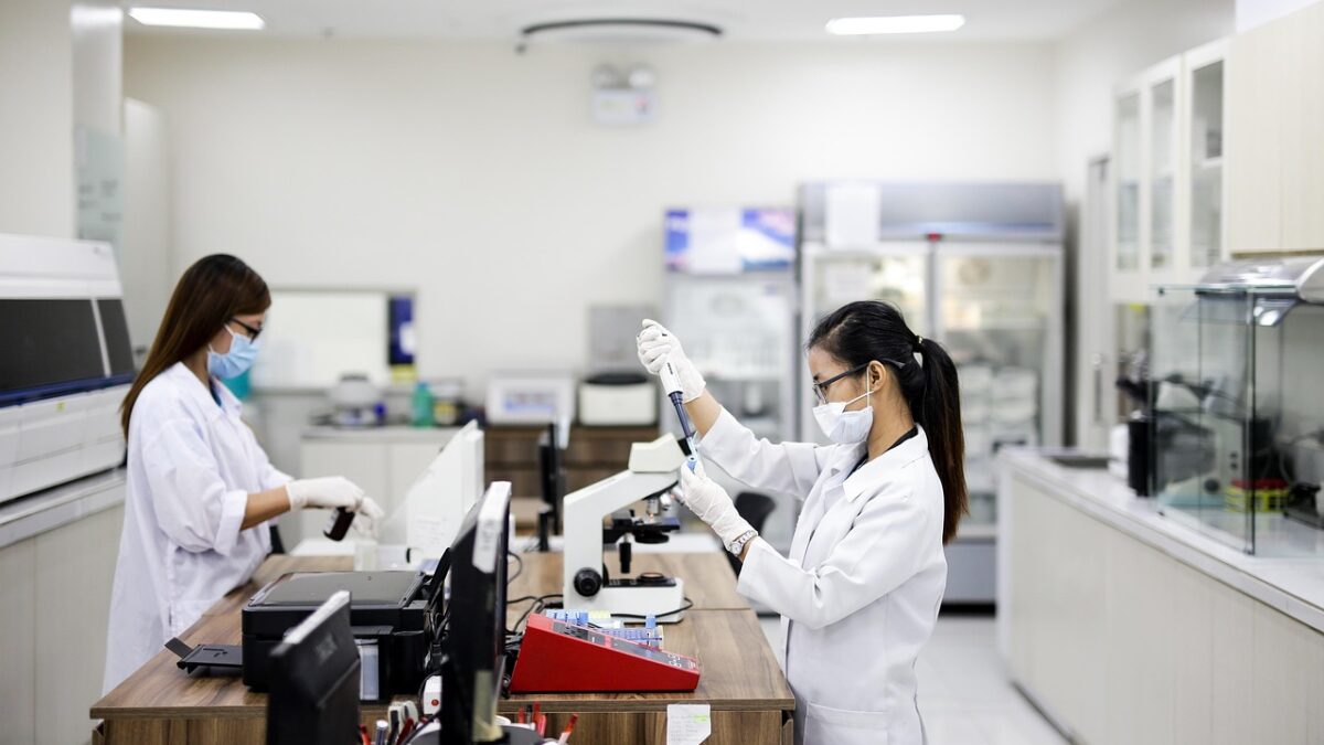Deux femmes en blouse blanche et masque travaillent dans un laboratoire clair : l'une manipule une pipette au-dessus d'un tube, l'autre est devant un ordinateur. Microscope, imprimante et réfrigérateur sont visibles en arrière-plan.