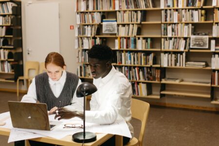 Two students studying together with a laptop in a library. Collaborative learning and academic research.