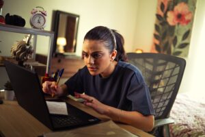 A concentrated woman working intensely on a laptop, taking notes at a home office desk.