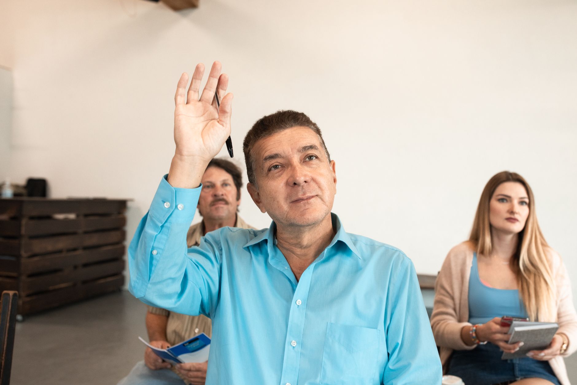 Man in a blue shirt raising his hand, with two seated people holding notebooks behind him.