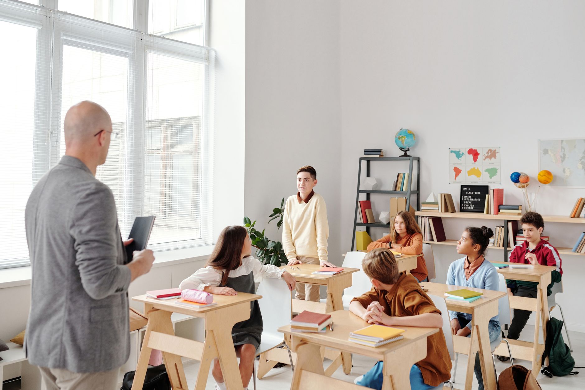 A male teacher with a folder talks to students seated at desks in a bright classroom.