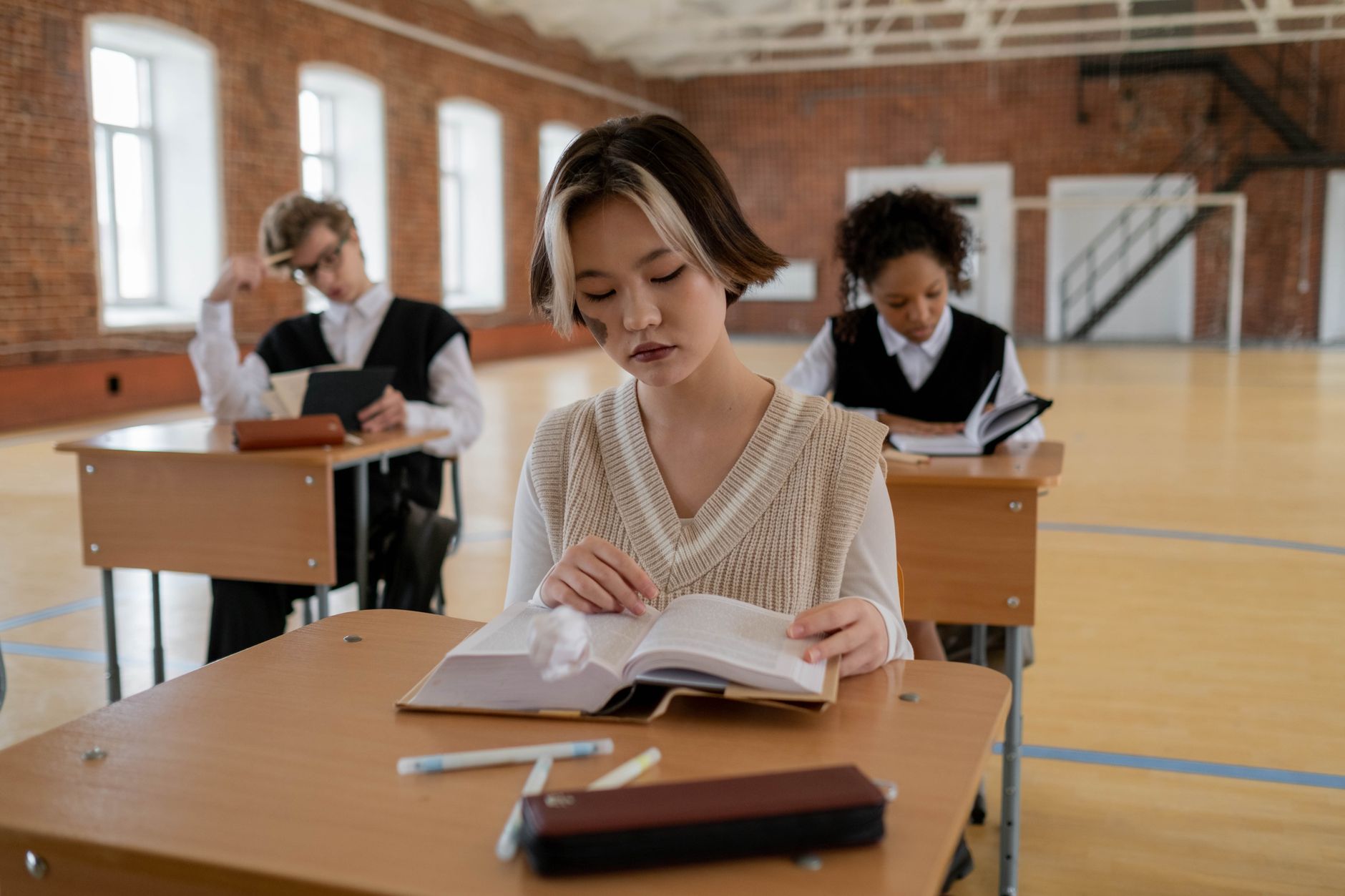 A student in a knit vest reads an open book at a desk in a brick hall, with two other students seated at desks behind.